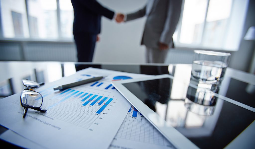 Image of eyeglasses, glass of water, touchpad and financial documents at workplace with businessmen handshaking on background